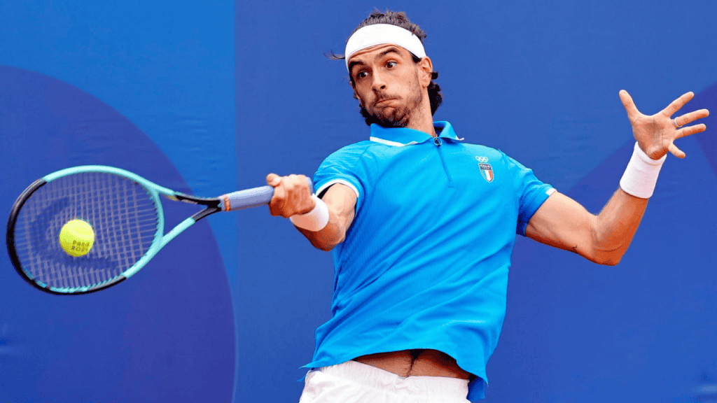 tennisser met baard en hoofdband die een bal met een forehand retourneert tegen een blauwe achtergrond